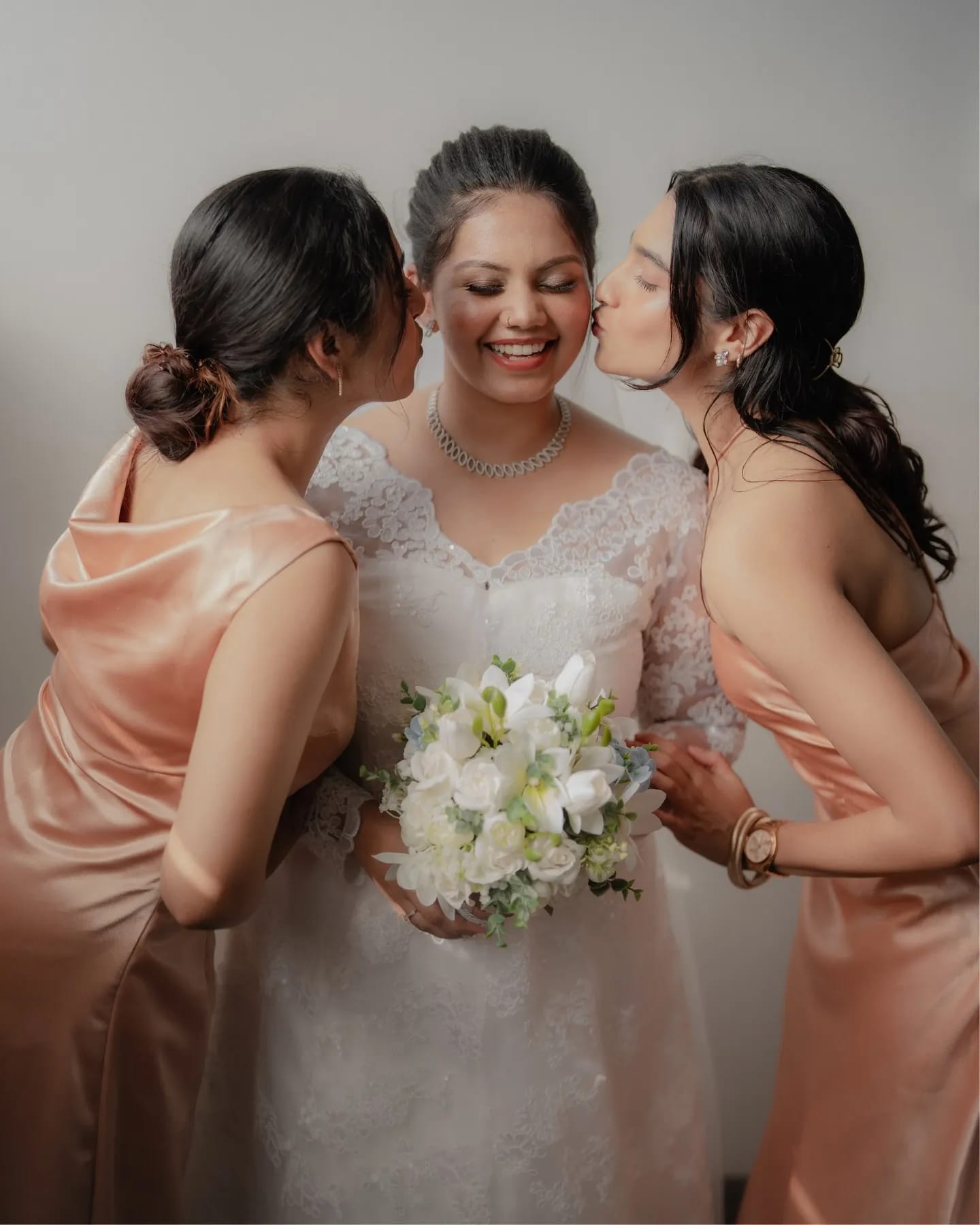 Two bridesmaids kissing a smiling bride on her cheeks