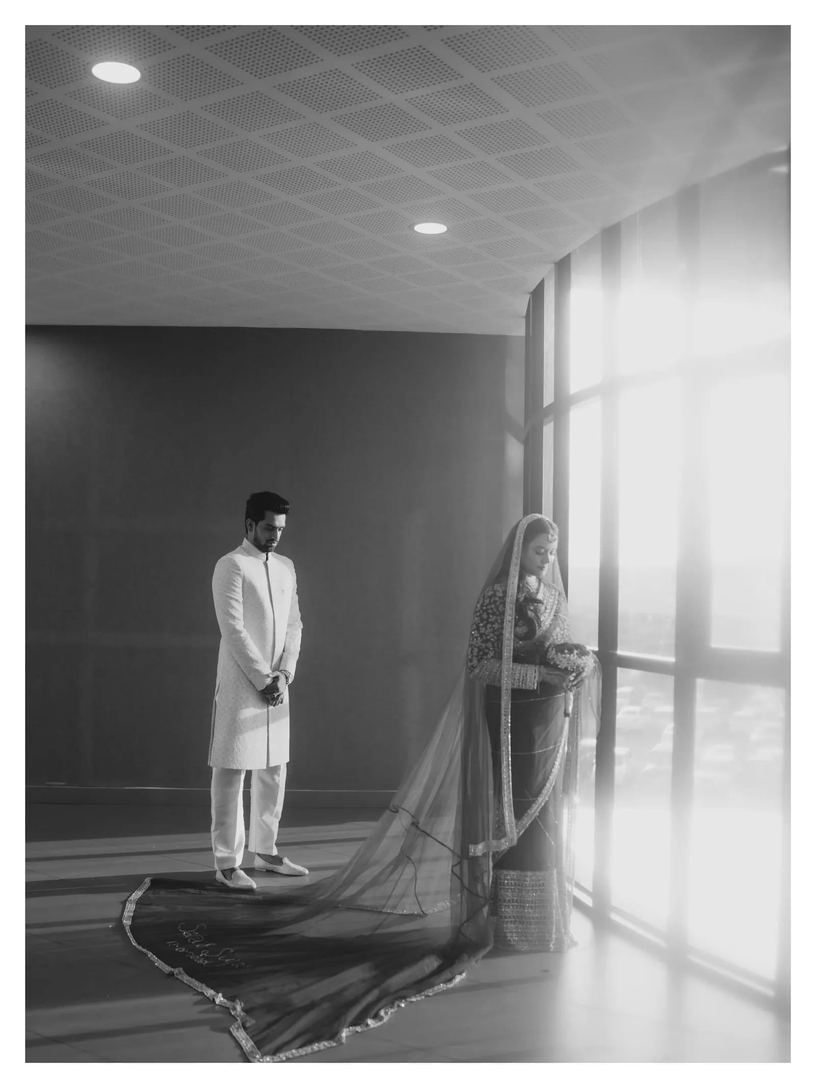 Black and white portrait of a couple beside a tall window, with the bridal veil trailing across the floor