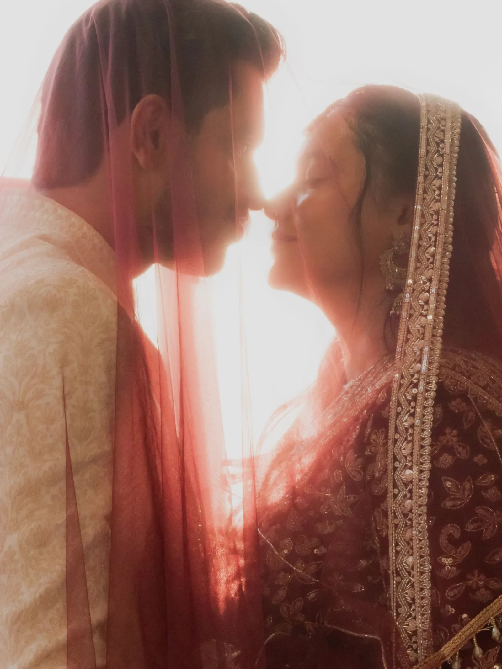 Bride and groom sharing a quiet moment beneath a sunlit red veil